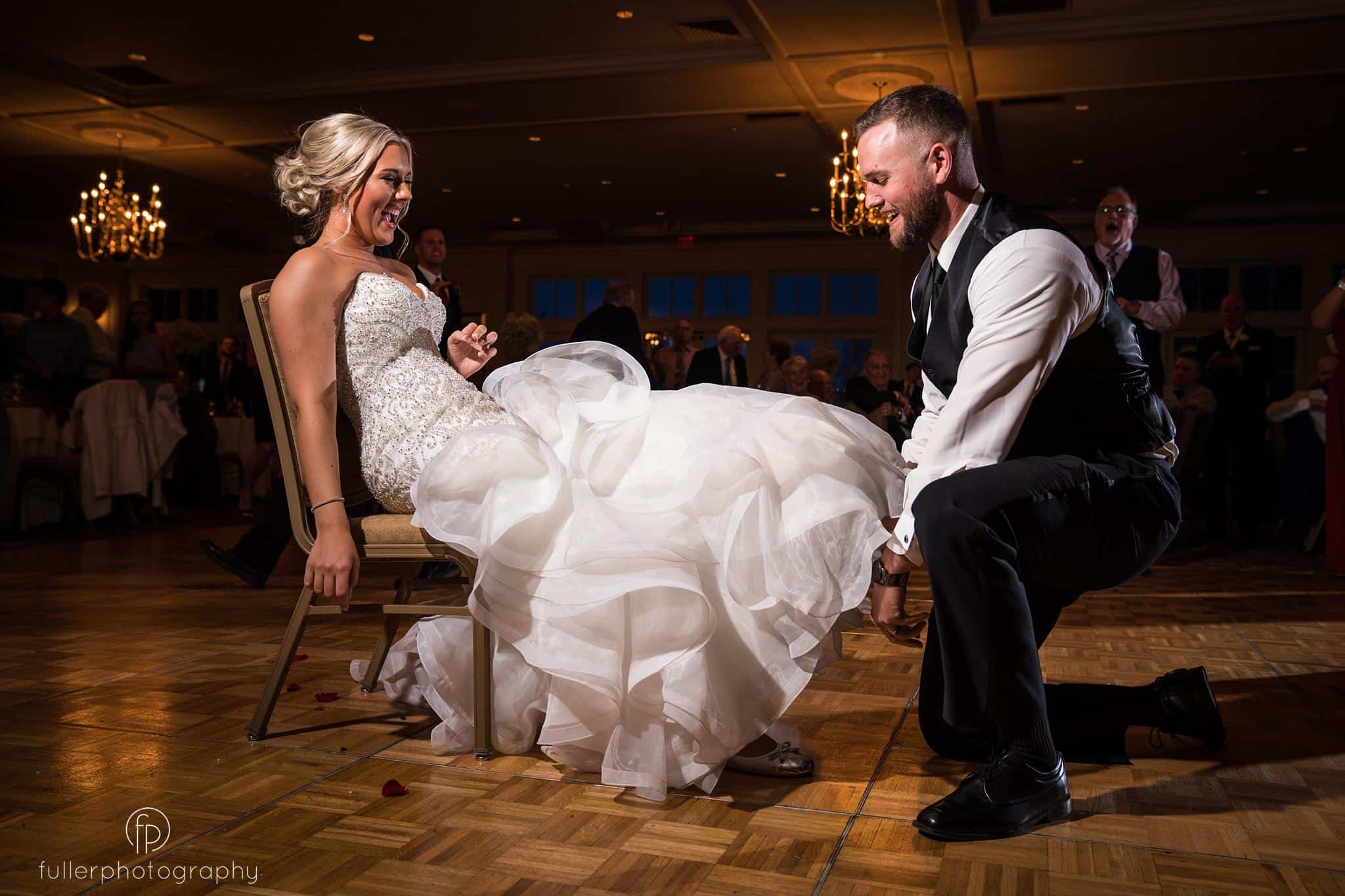 The bride and groom laughing as the groom takes off the garter from her leg on the ballroom floor in front of the wedding guests
